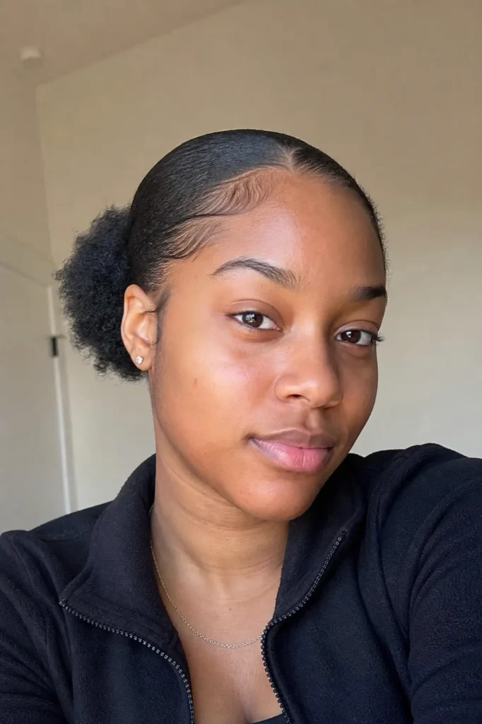 a young Black woman with a low puff hairstyle and smooth, laid edges. She wears minimal makeup and small stud earrings,