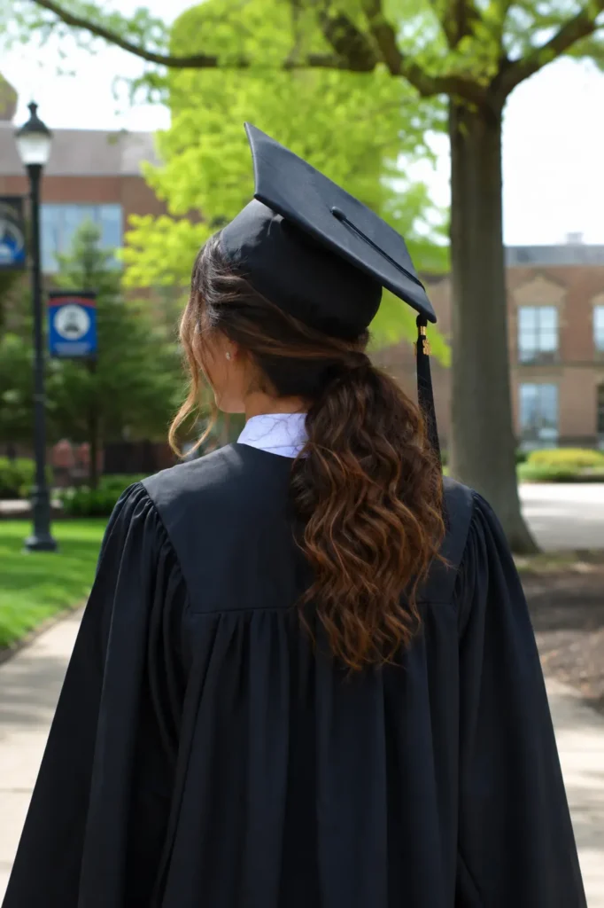 Low textured ponytail graduation hairstyle viewed from the back on a real young woman wearing a graduation gown and cap slightly tilted back, showing soft volume and natural movement in the ponytail. Realistic campus background with daylight