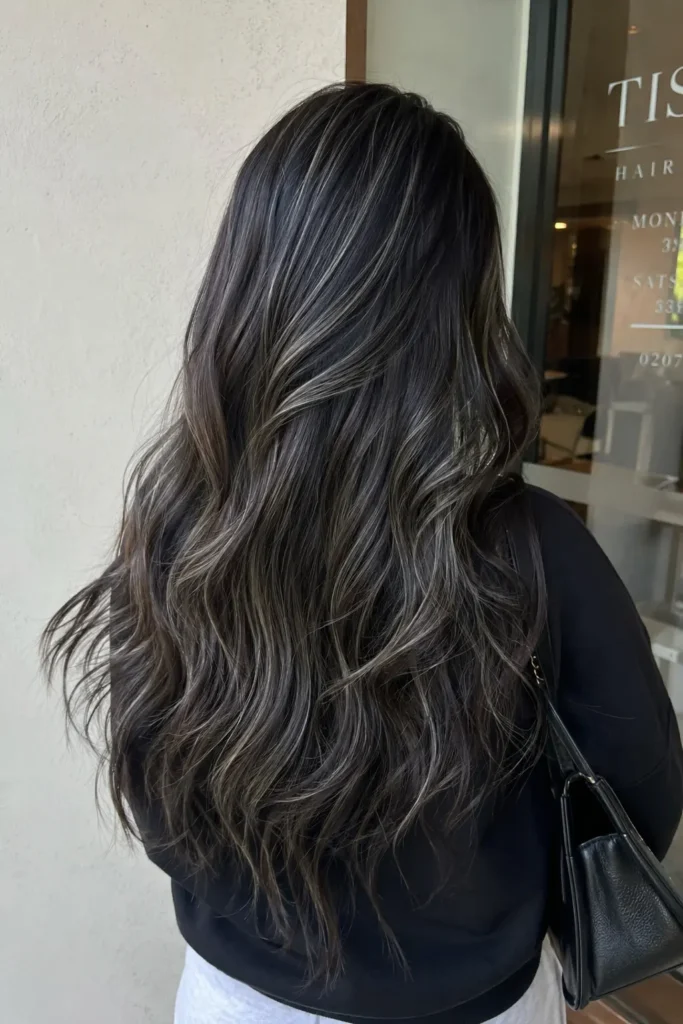 Back view of a woman with long dark hair styled in soft waves and cool-toned ash babylights, standing near a salon entrance in natural light.