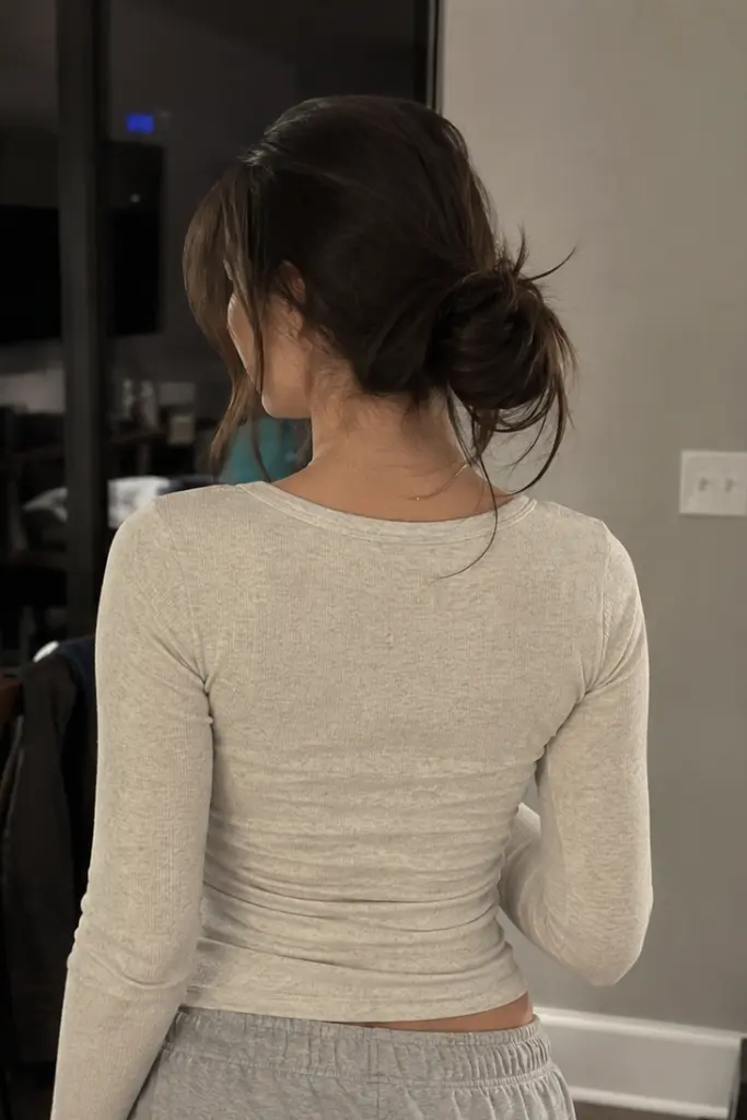 Back view of a real girl with a messy low bun hairstyle, soft loose strands framing her face, wearing a cozy neutral outfit indoors with warm lighting and realistic hair texture.