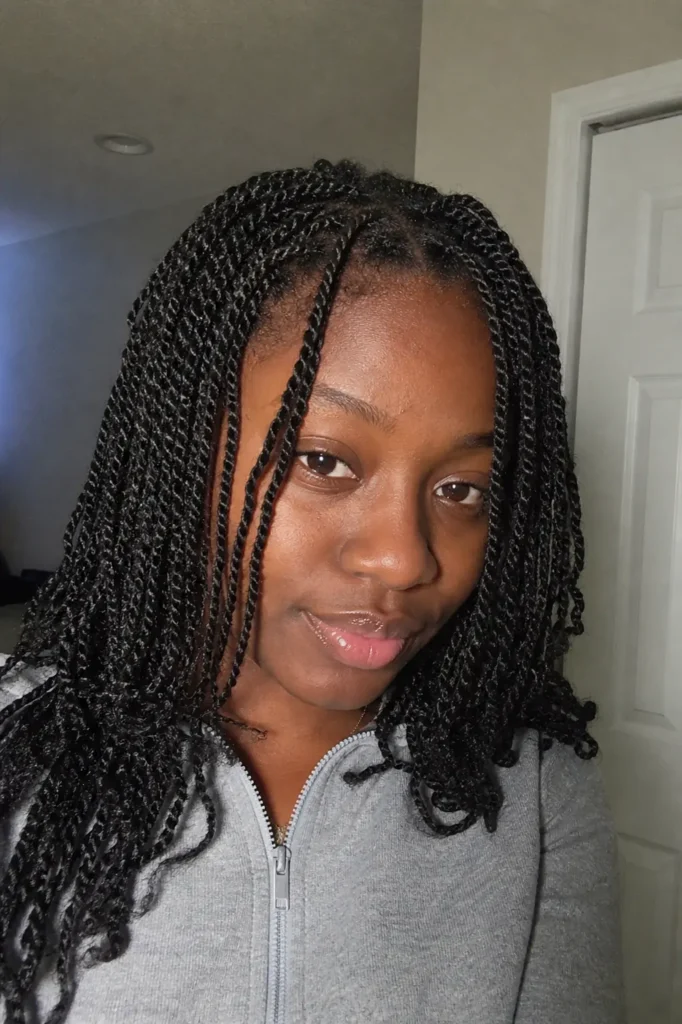 a young Black woman with small, neat mini twists, photographed indoors under soft lighting. She has a natural, relaxed expression, minimal makeup, and smooth skin