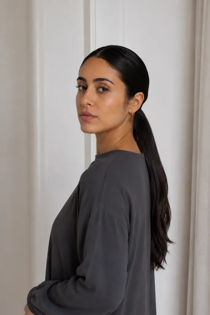 a woman in her late 20s with a minimalist slicked low ponytail hairstyle, featuring glossy sleek roots and a polished finish. She wears modest long-sleeve dark gray clothing