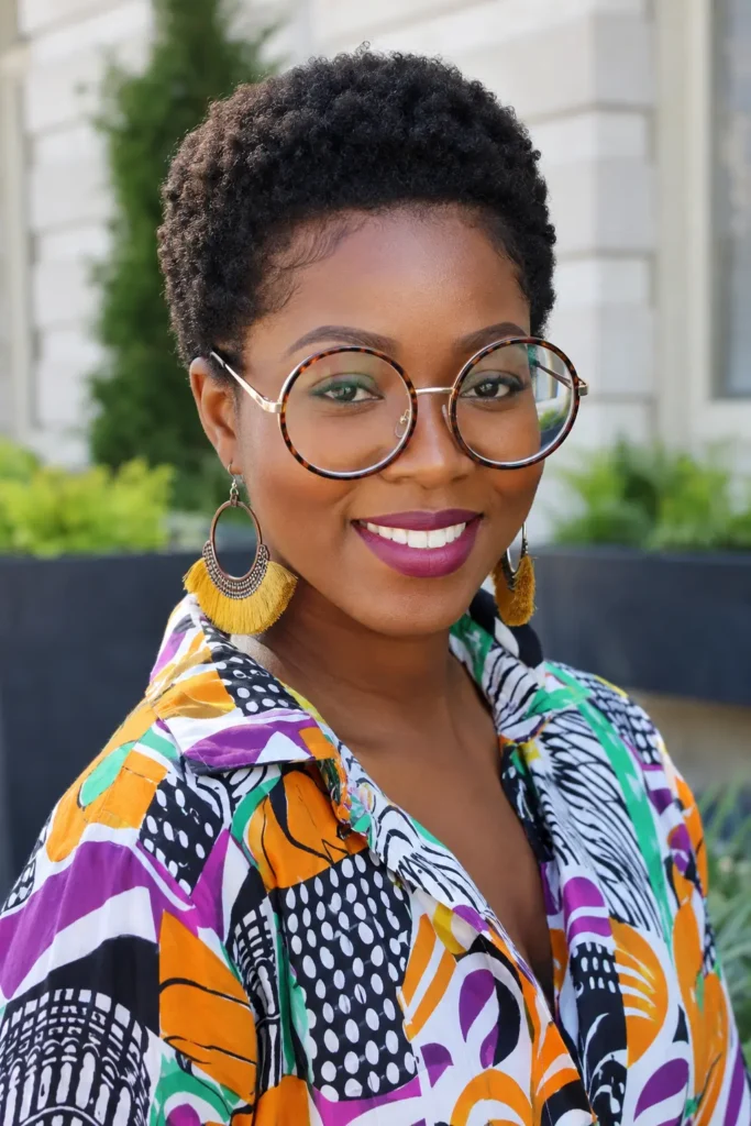 A smiling Black woman in her late 20s stands outdoors in bright natural light, wearing oversized round glasses and a colorful patterned blouse. She has a neatly shaped natural Afro pixie cut with defined edges and rich dark brown coily hair. Large tassel earrings and her confident expression add to the vibrant, authentic portrait style