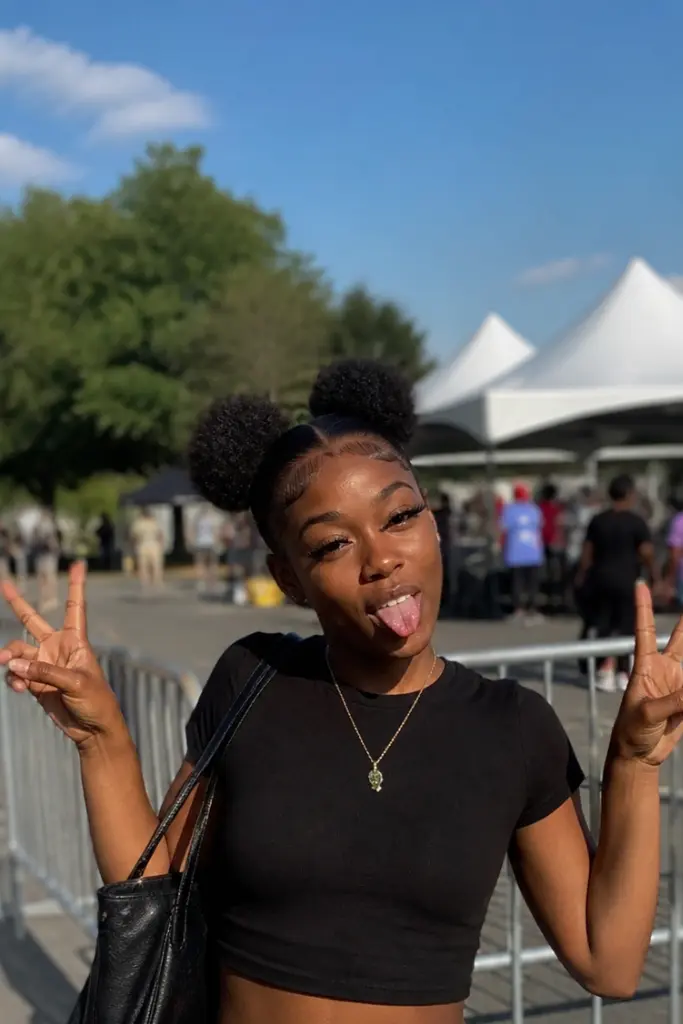 A young Black woman with natural space buns hairstyle poses playfully outdoors in bright daylight, sticking out her tongue and holding up peace signs with both hands. She wears a fitted black crop top, light blue high-waisted jeans, and a small necklace, with a black shoulder bag on one side.