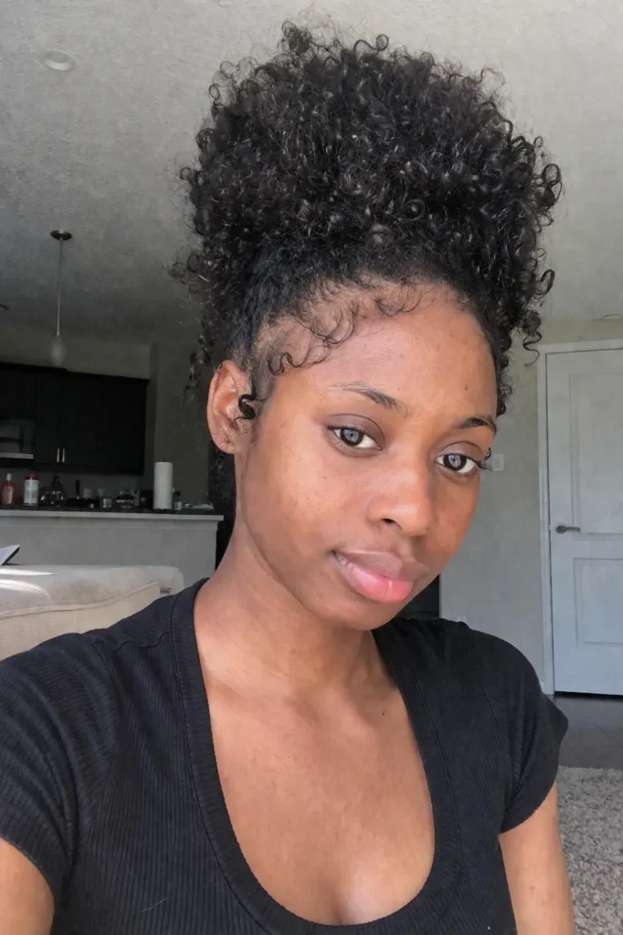 a young Black woman with a pineapple updo—voluminous natural curls gathered on top—wearing a black top, softly lit by natural daylight in a relaxed home setting