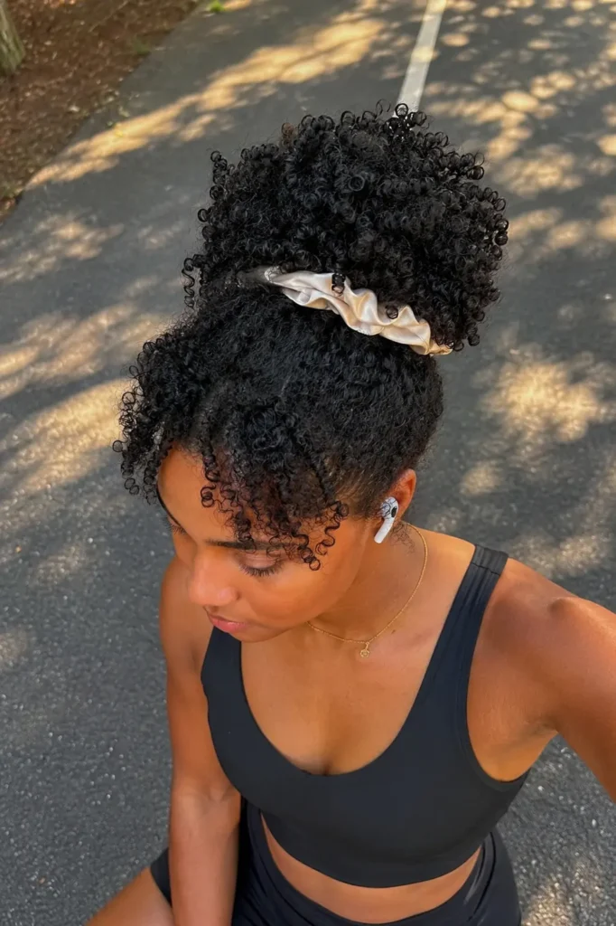 Young woman with defined 4A natural curls styled in a high pineapple updo secured with a satin scrunchie, wearing black running gear outdoors