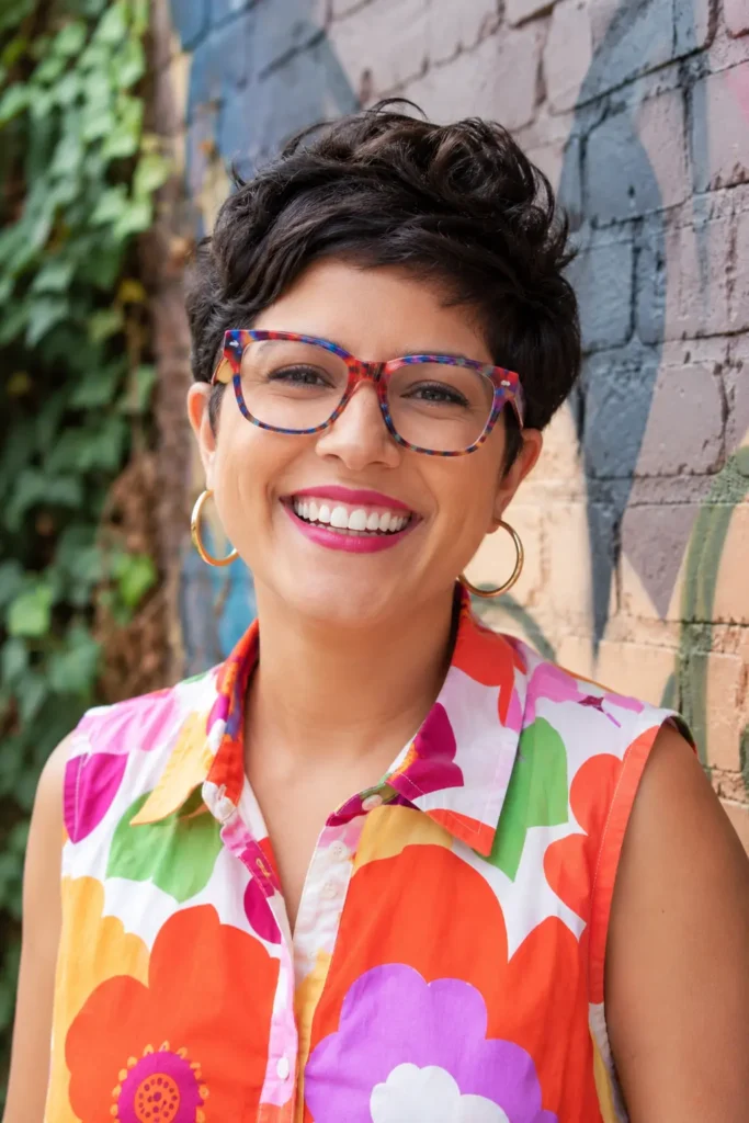 A joyful Latina woman in her early 30s stands outdoors in bright natural light, smiling warmly at the camera. She has a thick dark brown pixie haircut with soft natural volume and wears bold multicolored acetate eyeglass frames