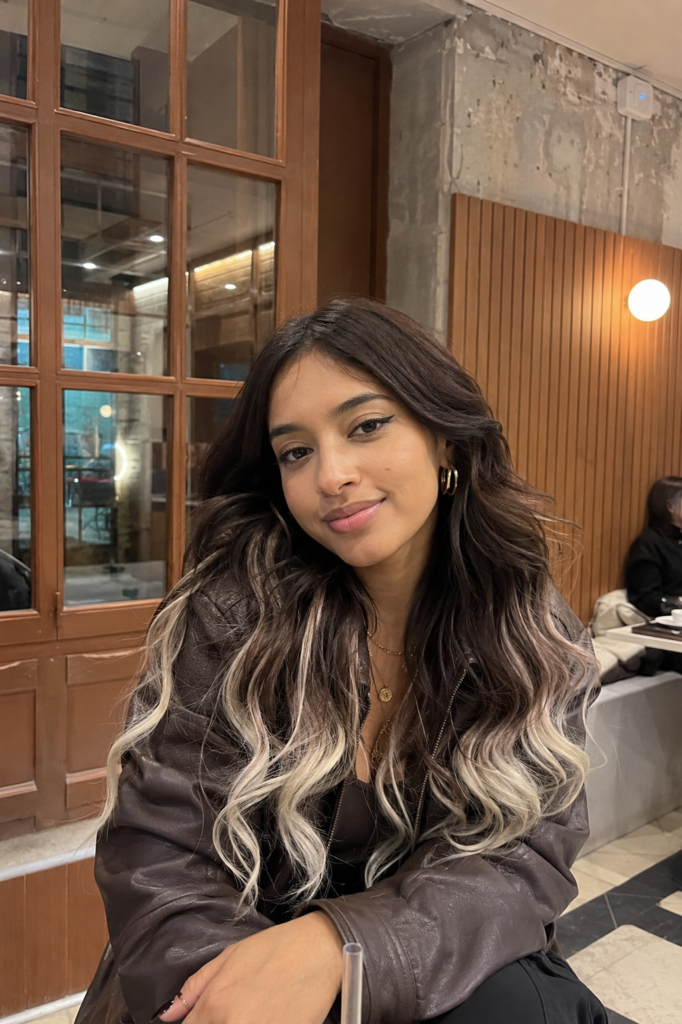 a young woman with warm beige skin sitting casually in a trendy café, wearing a dark espresso brown deep wave wig with icy platinum blonde tips, layered gold jewelry, and a brown leather jacket