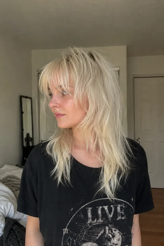 young woman with a platinum blonde wolf cut hairstyle featuring shaggy textured layers, icy blonde tones, messy crown volume, and wispy bangs. She is wearing an edgy casual black graphic T-shirt in a softly lit indoor bedroom setting