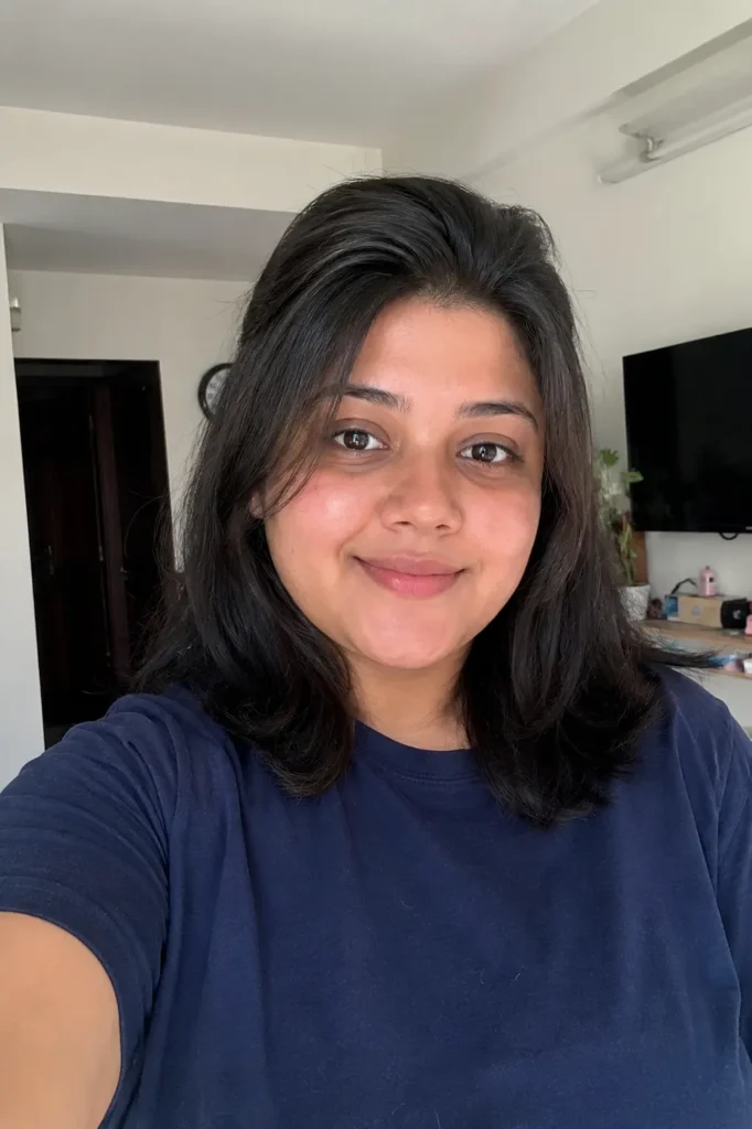 real young woman with a round face shape and shoulder-length haircut featuring soft crown volume, standing indoors in natural daylight.