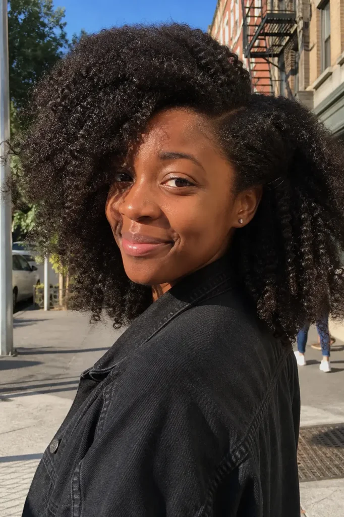 Young Black woman with a side-part afro and full, textured curls, smiling softly in natural sunlight on a city street with an urban background.