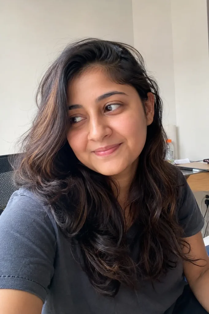 A real young woman with a round face shape and long loose waves styled in a deep side part, sitting indoors in soft natural light