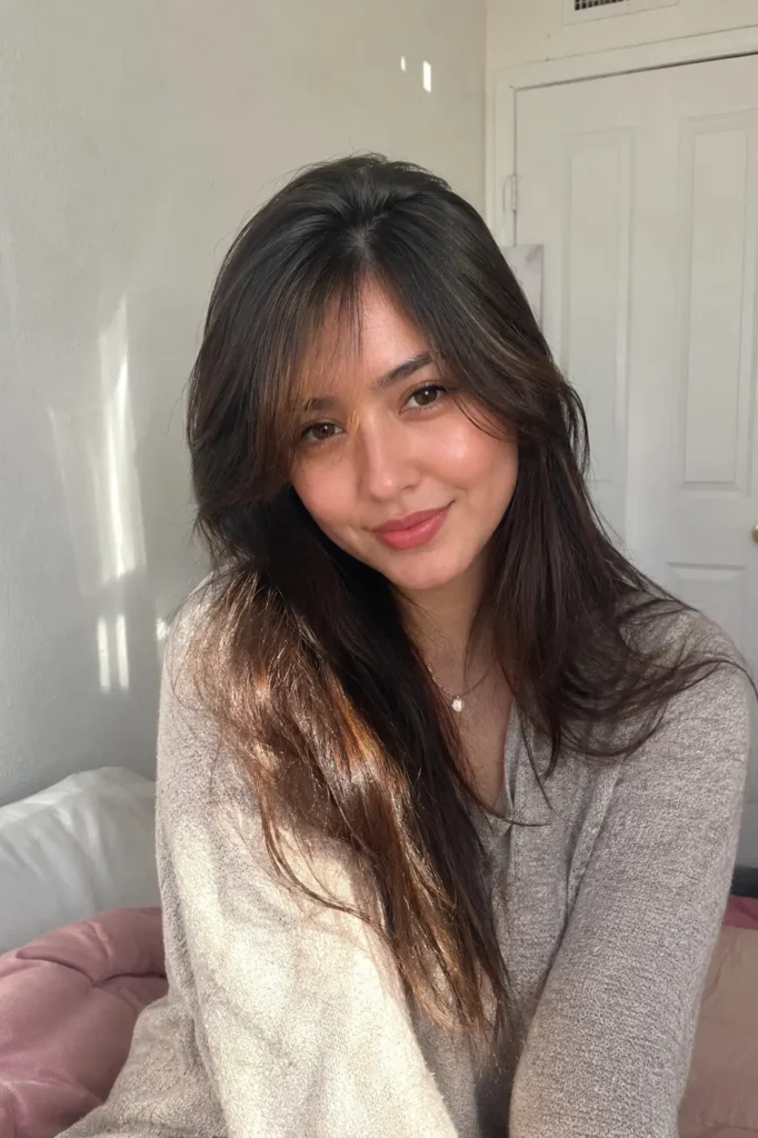 a young woman with a heart-shaped face and long dark brown hair styled with soft side-swept bangs, sitting indoors in cozy natural daylight. She wears a light beige sweater