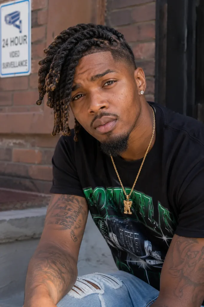 a Black man with a side-swept barrel twist loc hairstyle and a clean taper fade. He has natural skin texture, groomed facial hair, and authentic facial features. Wearing a black graphic T-shirt, ripped light-wash jeans, a gold chain