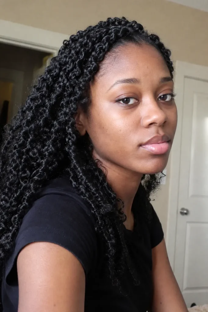 a young Black woman with side twists styled into loose, defined curls, softly lit in a natural indoor setting. She wears a simple black top