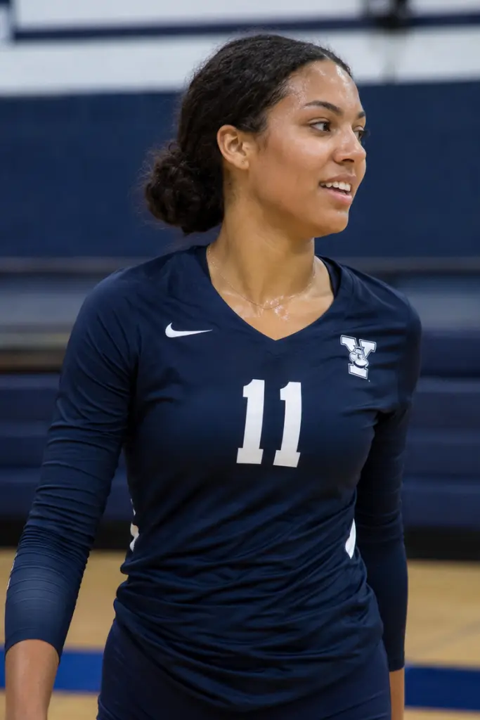 College-age volleyball player with natural curly hair in a low bun, wearing a dark athletic uniform in an indoor gym