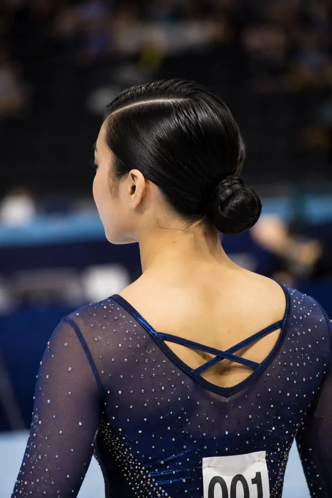 Young Asian gymnast with sleek straight black hair styled in a polished side-parted low bun, viewed from a slightly elevated back angle, wearing a sparkling navy gymnastics competition outfit in an indoor arena.
