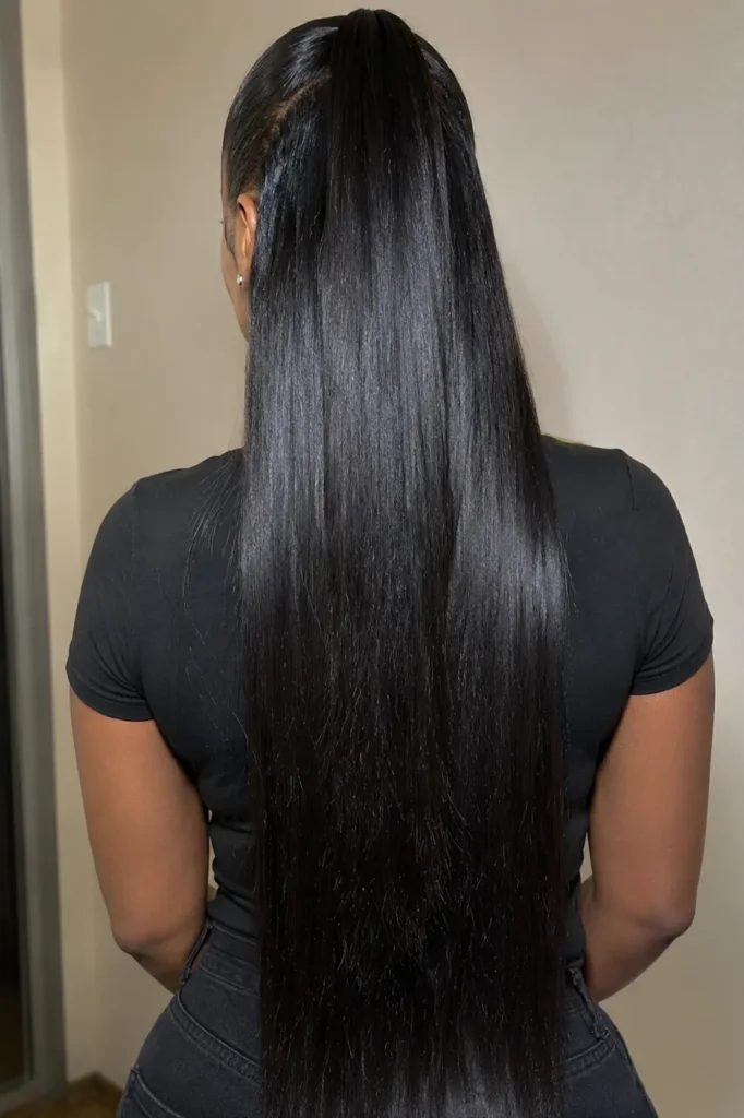 Back view of a real Black woman with a sleek straight half up half down ponytail hairstyle. Glossy silky black hair flows smoothly down her back with a polished high ponytail top section