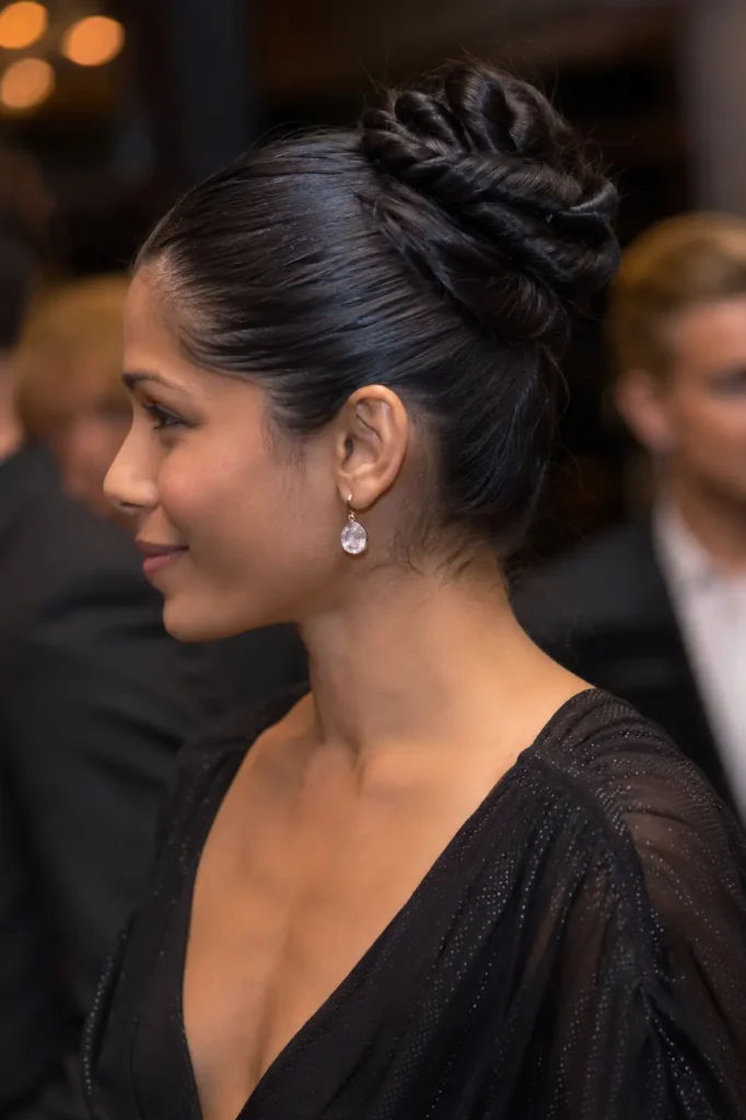 a woman in her late 20s with a sleek front-twisted bun hairstyle featuring glossy, sculpted twists. She wears an elegant black dress with subtle shimmer details and delicate drop earrings.