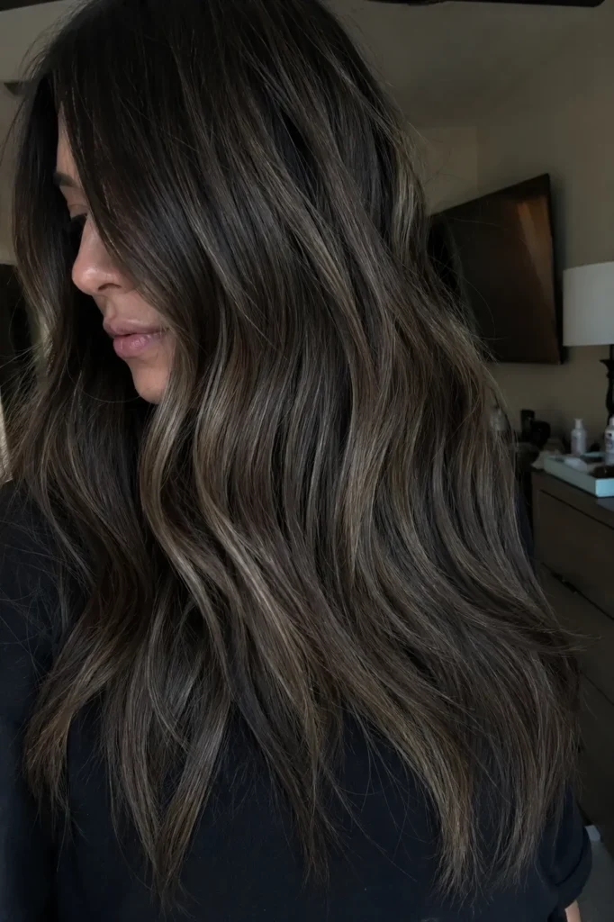 a woman indoors with dark espresso hair featuring smoky ash babylights, styled in soft layered waves.