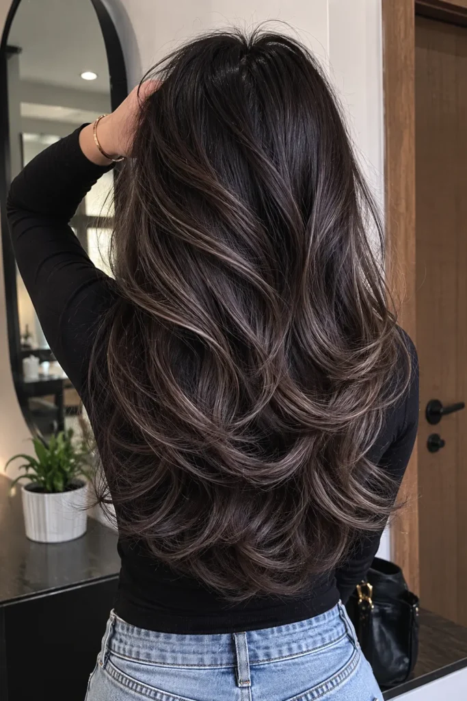 Back view of a real young woman with dark hair featuring smoky gray ash babylights, styled in soft layered waves with cool-toned dimension and texture