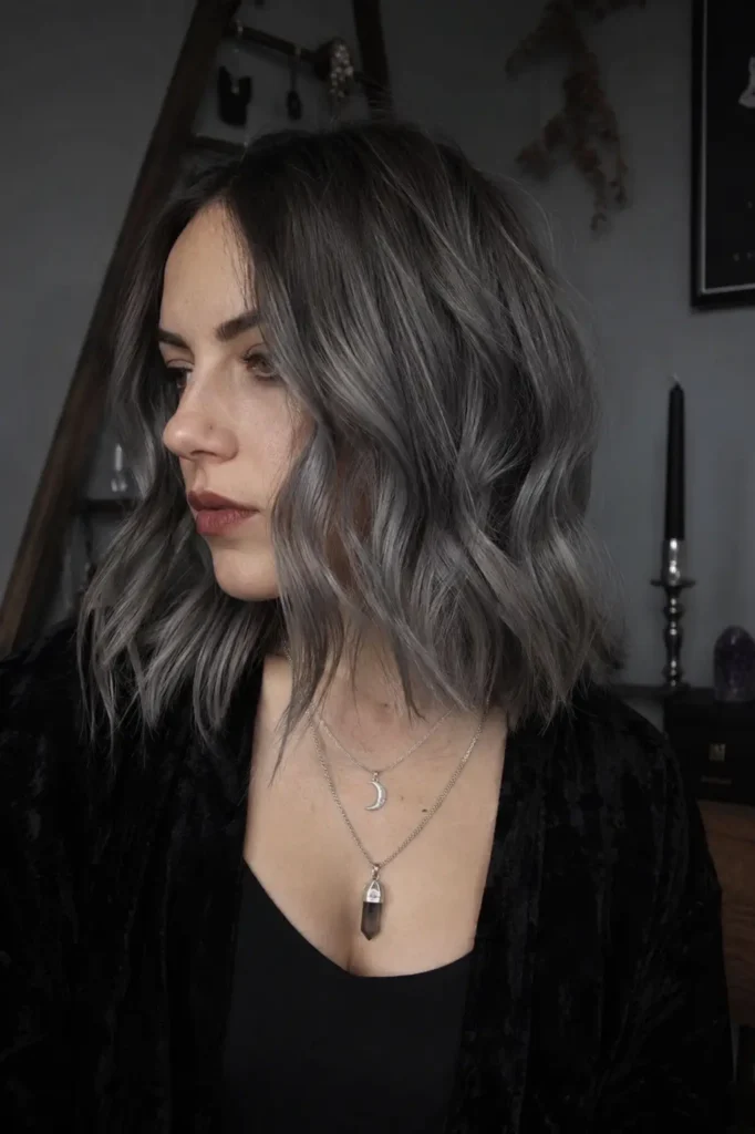 young woman with a smoky gray shoulder-length lob styled in soft loose waves, wearing layered silver jewelry and black witch-inspired clothing in moody soft lighting with a photorealistic modern mystical aesthetic.