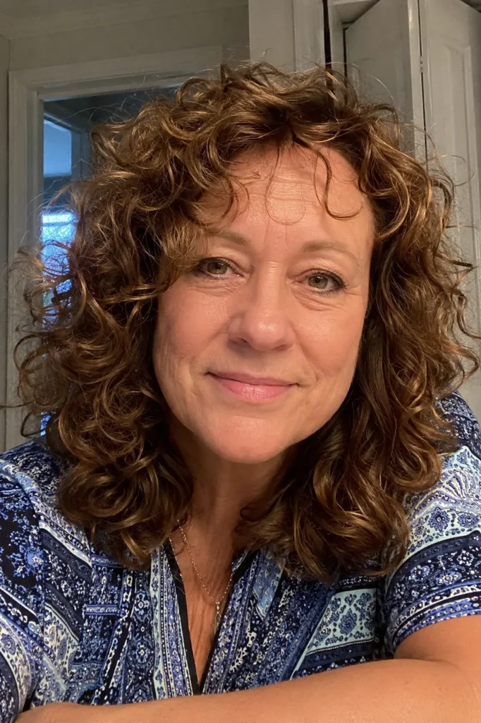 a woman around 50–60 years old with a soft curled mid-length hairstyle featuring natural volume and movement. She has authentic facial features, realistic skin texture,