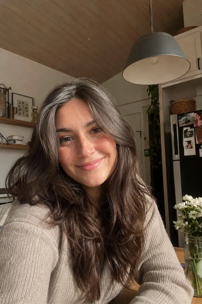 Real girl with soft face-framing thick wavy dark brown hair smiling naturally in a cozy indoor kitchen setting, warm daylight highlighting realistic facial features and natural makeup