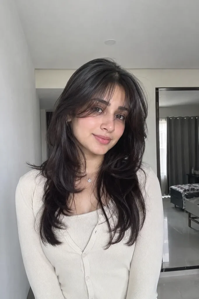 a soft feminine wolf cut hairstyle featuring blended feathered layers and airy curtain bangs, styled with glossy smooth texture and natural movement. She is wearing a modest elegant cream outfit