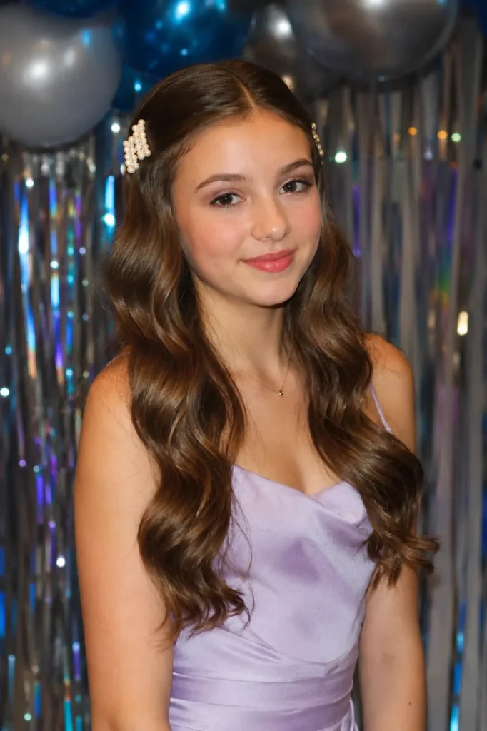 Teen girl with soft Hollywood waves styled in glossy, defined curls, accented by pearl hair clips. She wears a pastel lavender formal dress and subtle soft-glam makeup, posing at a school dance with a natural smile and realistic youthful features