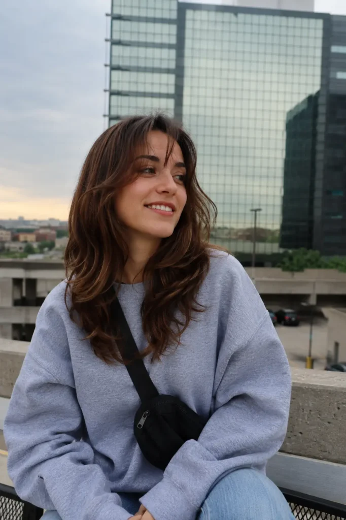 A real young woman with medium-length layered soft waves sitting outdoors in natural daylight, wearing a casual oversized sweatshirt and natural makeup,