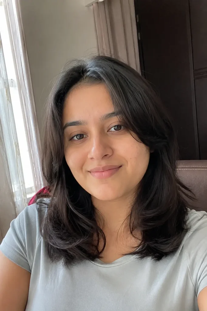 young woman with a square face shape and medium-length soft layered haircut sits indoors near a window in natural daylight. Her dark layered hair has a healthy realistic shine, framing her face with soft movement