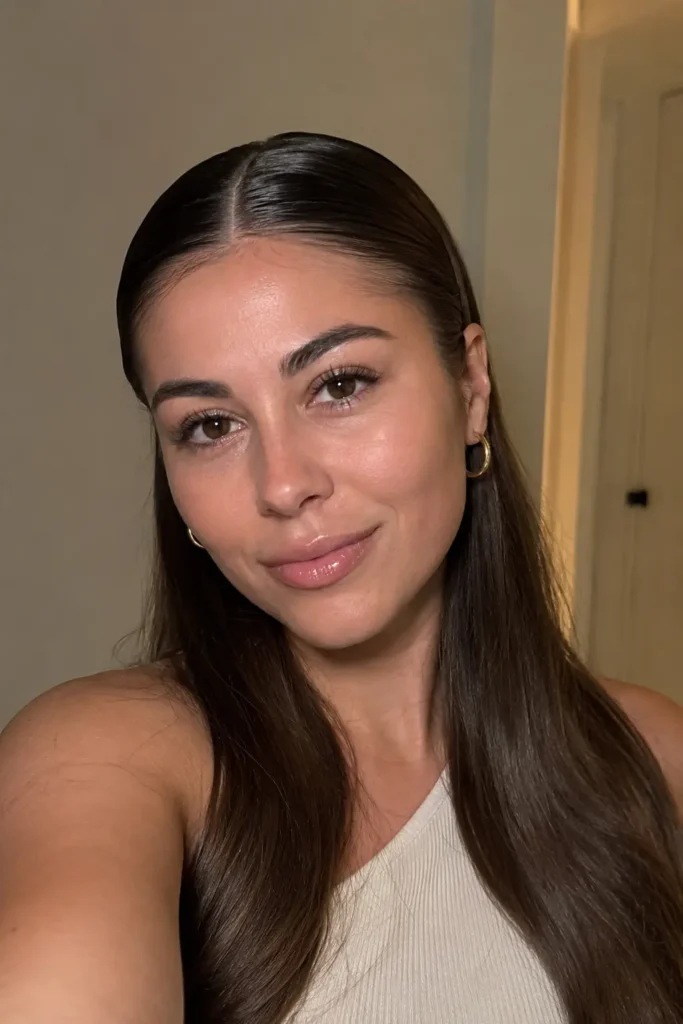 a young woman aged around 22–32 with authentic facial features, natural skin texture, and a calm relaxed expression. She has soft, loose brunette waves with subtle face-framing layers and a natural glossy finish