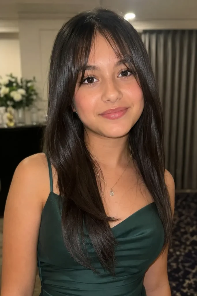 A realistic teenage girl with sleek, straight dark hair and soft curtain bangs poses in a modern formal dress. Her glossy, smooth hairstyle frames her face, highlighting natural facial features, soft glam makeup, and healthy skin texture.