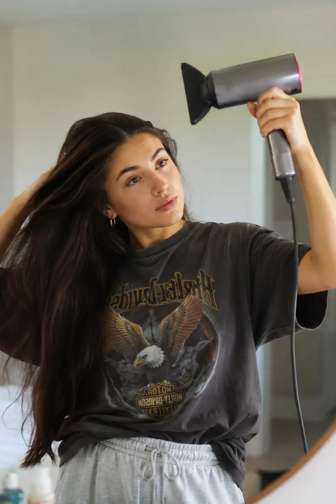 Young woman blow drying her thick dark hair while looking in a bedroom mirror, wearing casual loungewear in a cozy everyday setting with soft natural light and a relaxed Pinterest-inspired lifestyle aesthetic.