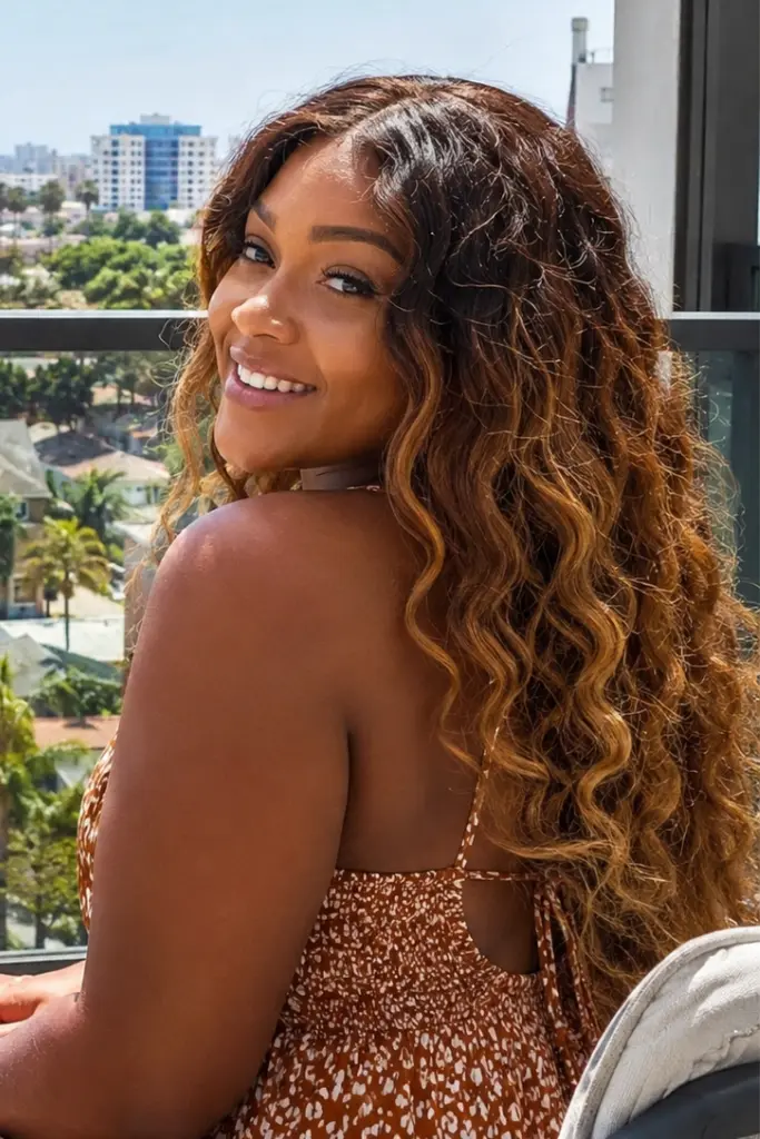 a smiling woman with warm medium brown skin sitting on a sunny balcony overlooking a cityscape, wearing a warm brown deep wave wig with golden blonde