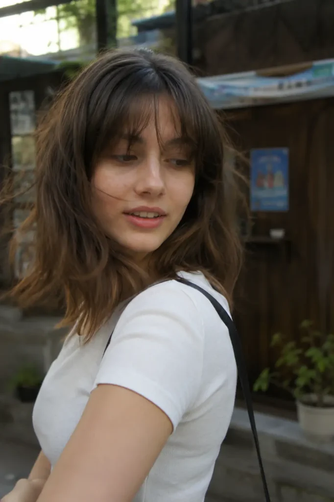 a real young woman with a tousled medium-length haircut and soft messy waves, wearing a casual white fitted top. She is captured in motion, glancing over her shoulder with loose strands framing her face