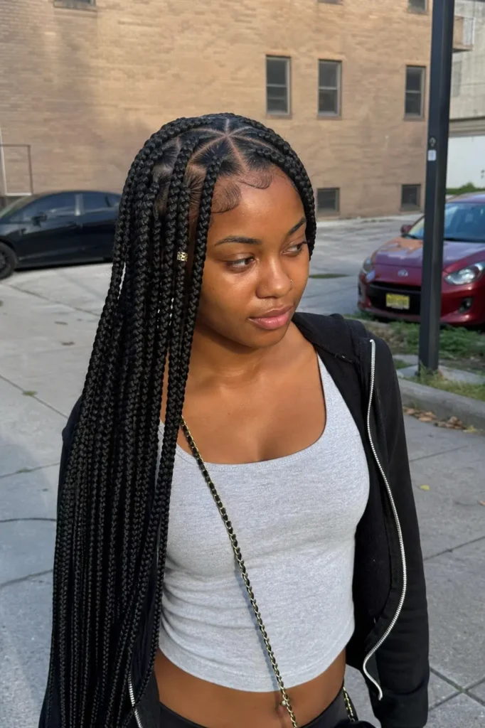 Real young Black woman with long triangle box braids and clean parts, wearing casual streetwear, standing outdoors in an urban setting with cars