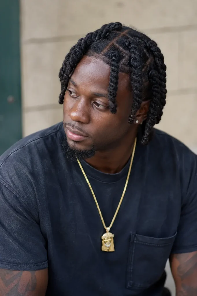 a Black man with a two-strand barrel twist loc hairstyle, featuring neatly sectioned twists and detailed texture. He has natural facial features, a groomed beard, and authentic skin texture. Wearing a dark casual T-shirt and gold pendant necklace