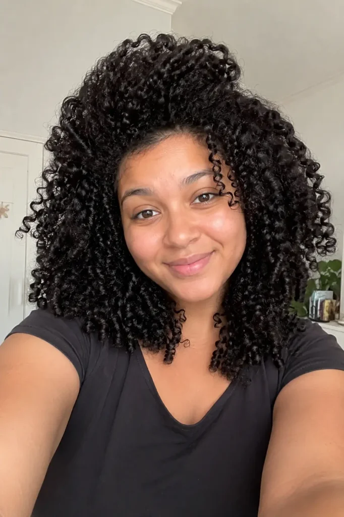 girl with a round face shape and voluminous curly hair featuring lifted roots and defined glossy curls, smiling in bright indoor lighting