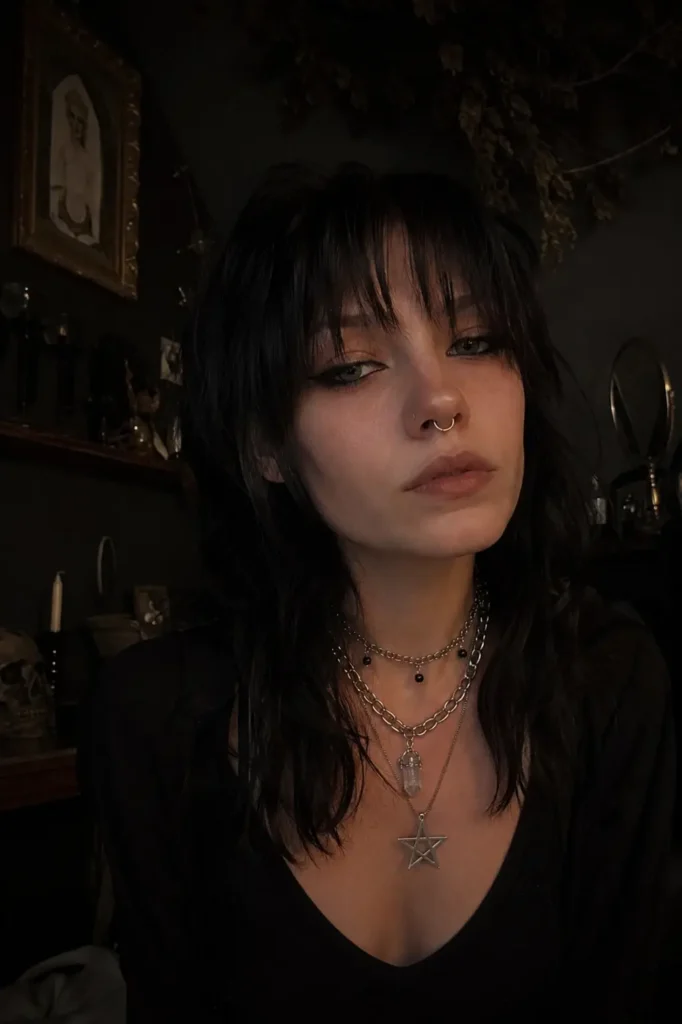 Real girl with a dark shag haircut and wispy bangs, wearing layered silver jewelry in a moody witchcore-inspired room with dim indoor lighting, alternative beauty aesthetic, photorealistic portrait with edgy gothic vibes.