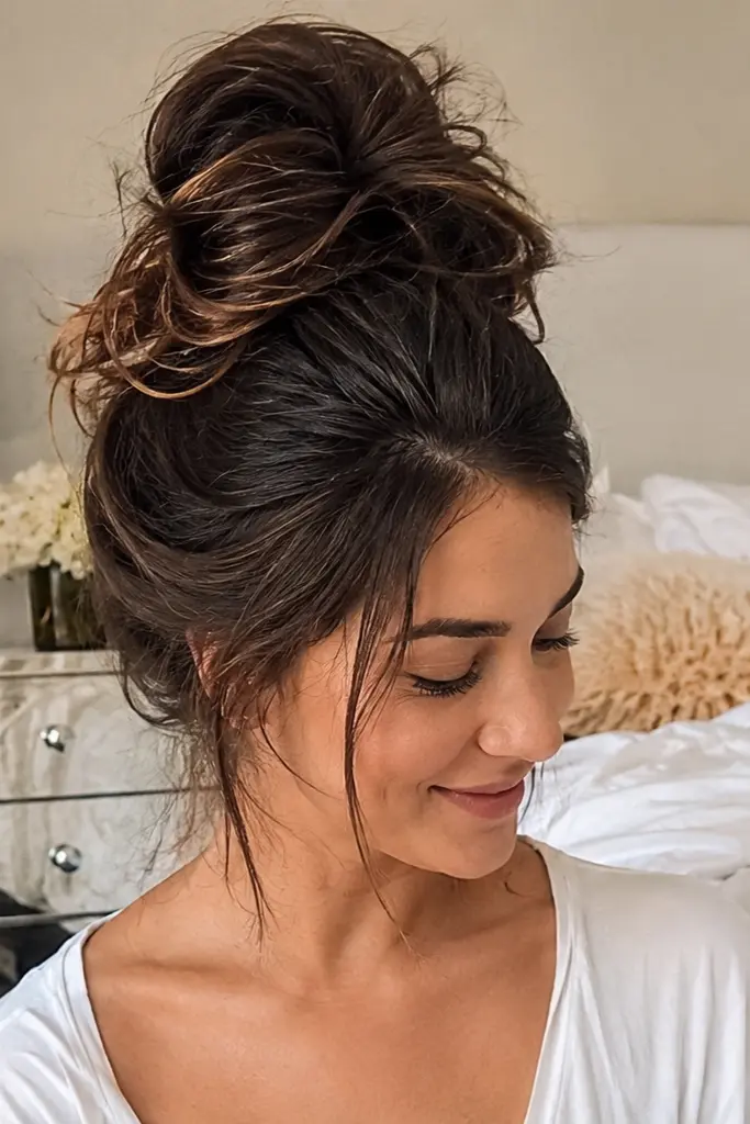 young woman in a cozy bedroom wearing a white top, with thick dark hair styled into an oversized messy top bun. She looks down with a soft, relaxed smile in warm natural daylight, with a neatly made bed, bedside lamp,