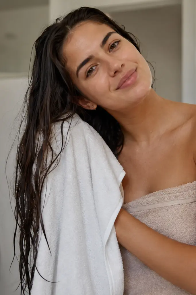 Real girl in a softly lit bathroom gently drying her long, thick wet hair with a white towel, wrapped in a beige bath towel,
