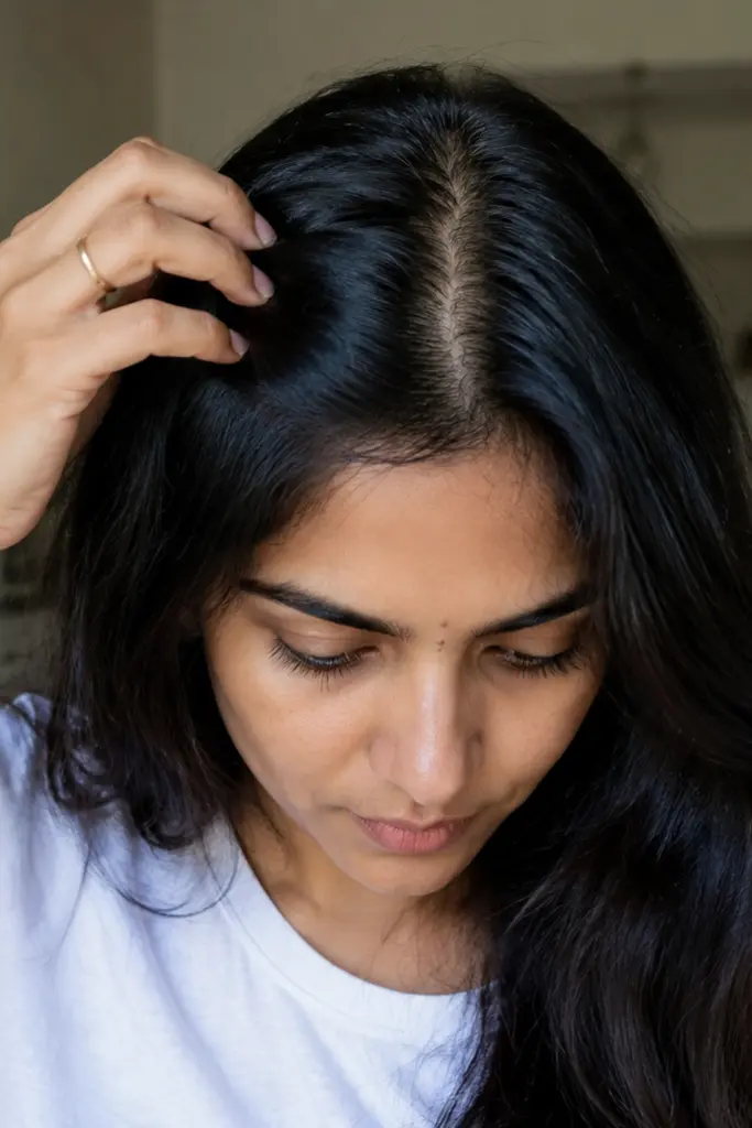 Woman indoors in natural daylight gently parting her thick dark hair, showing a clean center part with minimal scalp visibility. She wears a simple white top and looks down with a relaxed