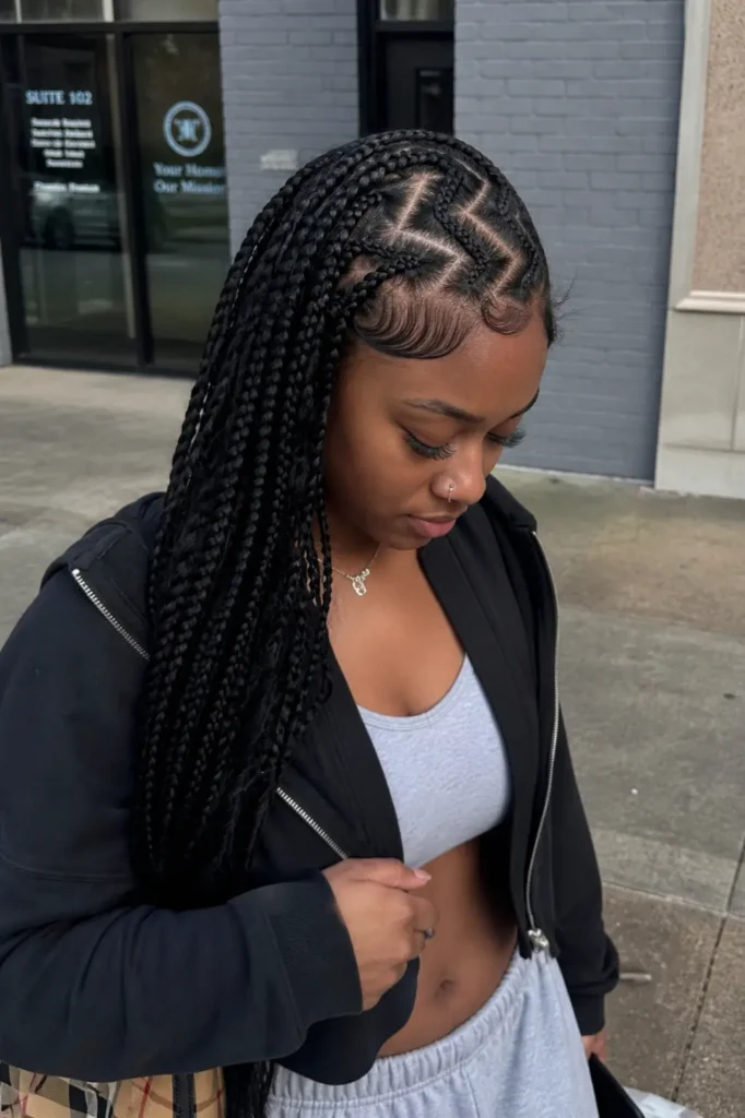 Young Black woman with zig-zag braids, casual outfit, standing outdoors in natural daylight with a candid urban vibe.
