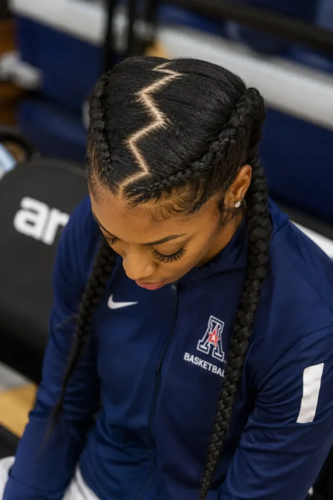 A young Black woman seated on a sports bench wearing a navy basketball warm-up jacket, photographed from a slightly elevated angle that highlights her intricate braided cornrows with a sharp zigzag scalp part, her head tilted downward in a calm