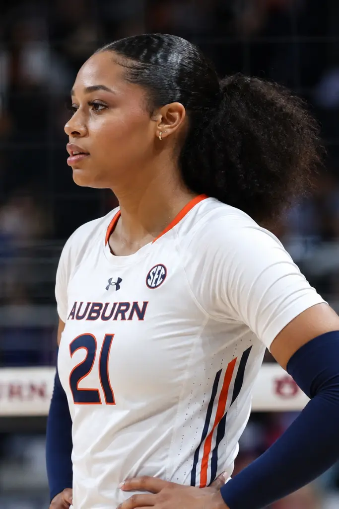 A focused young volleyball athlete stands on an indoor court wearing a white and navy volleyball uniform, featuring an athletic slick puff pony hairstyle with a sleek polished scalp and voluminous textured ponytail.