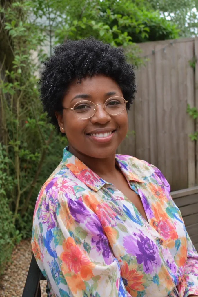 A smiling woman in her 30s with a voluminous natural curly pixie cut and dark coily 4C hair stands outdoors in soft natural light