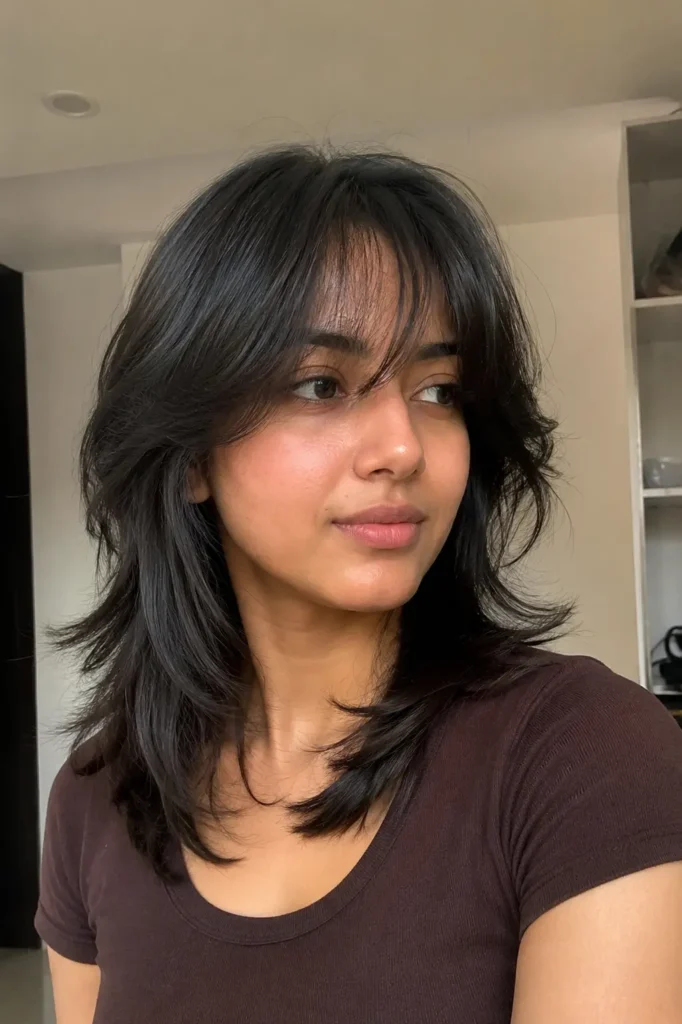 young woman with a shoulder-length wolf cut hairstyle featuring feathered textured layers and a soft side fringe. Her dark hair has natural shine and movement, styled with airy volume indoors under soft daylight