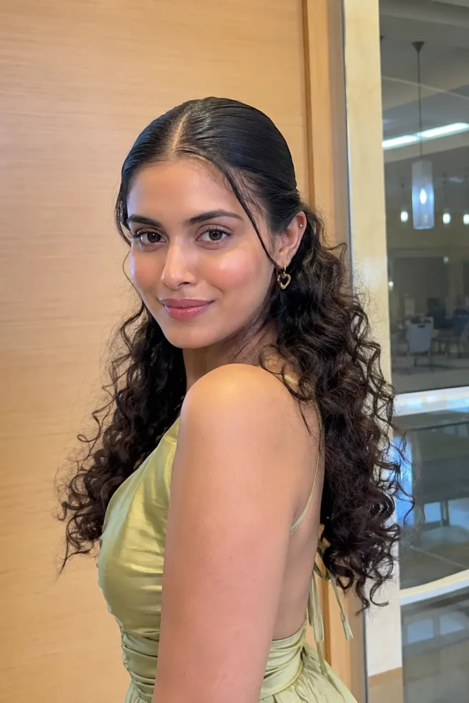 Young woman with slicked front hairstyle and defined loose curls, glowing natural skin, and a soft feminine expression wearing an elegant satin dress in a vertical indoor portrait.
